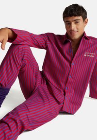 Mladý muž sedící na podlaze, oblečený v červeno-fialové pruhované košili na knoflíky a odpovídajících kalhotách s nápisem "Benetton" na hrudi.