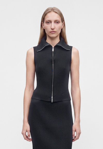 Femme aux longs cheveux blonds portant un haut sans manches noir côtelé zippé et une jupe assortie, debout devant un fond clair uni.