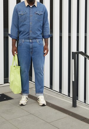 Chaqueta de denim con un collar marrón, jeans a juego y una bolsa de tote verde claro. Usado con zapatillas crema y negras sobre una superficie texturizada.