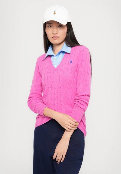 Roza pletena jopica z V-izrezom in rebrastimi manšeti, oblečena čez svetlo modro srajco s ovratnikom, v kombinaciji s temno modrimi hlačami.