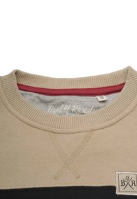 Beige sweatshirt med ribbat krage, med röd innerfoder och svart rand. Inkluderar en logopatch och X-sömnadsdetaljer.