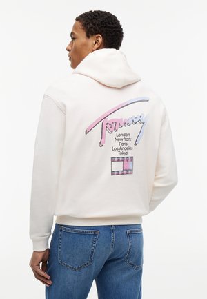 Άνδρας που φοράει λευκό φούτερ Tommy με παστέλ λογότυπο και ονόματα πόλεων, συνδυασμένο με μπλε τζιν, με την πλάτη προς την κάμερα.