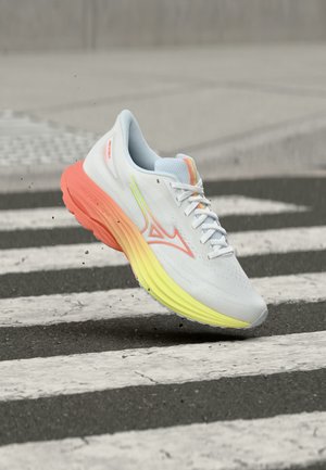 Witte hardloopschoen met oranje en gele verloopzool hangend boven een zwart-wit gestreept zebrapad.