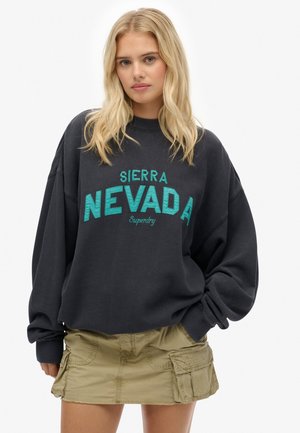 Zwarte oversized sweatshirt met de tekst "SIERRA NEVADA" in teal, gecombineerd met een khaki cargorok met zijzakken en een gemëleerde stof.