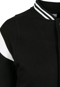 Schwarze Sportjacke mit weißen Akzenten, gerippter Textur, gestreiftem Kragen und Druckknopfverschluss. Nahaufnahme des Schulter- und Kragenbereichs.