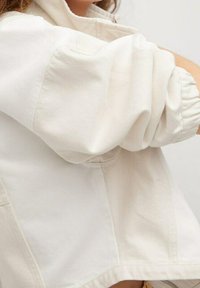 Krémová bavlněná bunda s volným střihem, která má nabírané rukávy a viditelné švy. Minimalistický design, bez kovových doplňků.