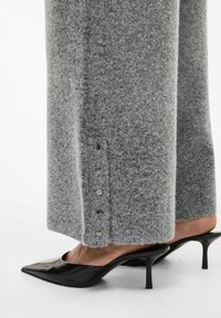 Szare spodnie z wełnianego materiału o szerokich nogawkach, z zapięciami na napy wzdłuż dolnej krawędzi, w połączeniu z czarnymi, lakierowanymi klapkami na obcasie.