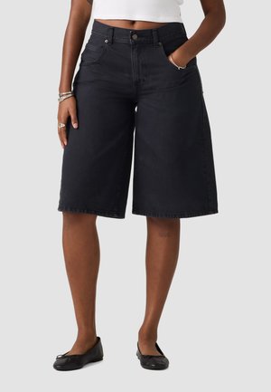 Personne portant un short large noir au niveau des genoux, un top court blanc, des ballerines noires et des bijoux argentés à un poignet et sur deux doigts.