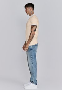 Beige aansluitend t-shirt met lichtblauwe kleuraccenten bij de kraag, gecombineerd met vervaagde blauwe jeans en witte sneakers, weergegeven vanuit de zijkant.