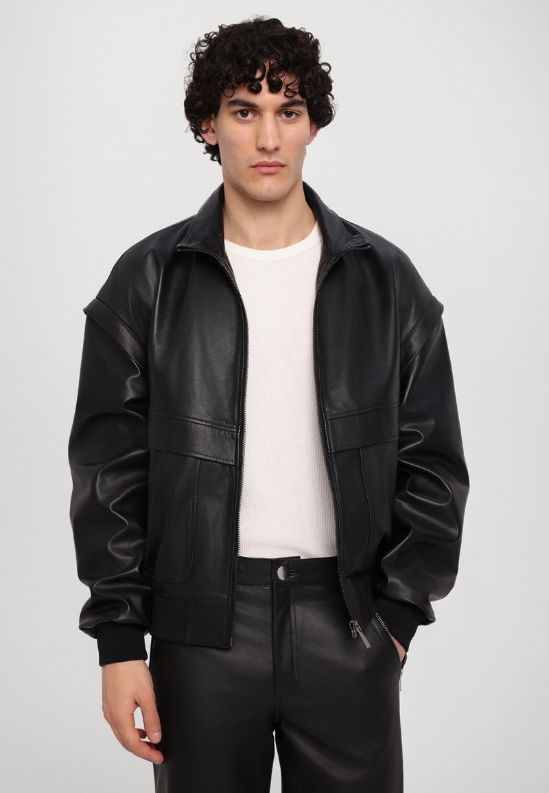 Black leather jacket with a zip front, ribbed cuffs, and two front pockets. Worn over a white fitted shirt and black pants.