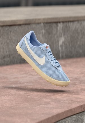 Ανοιχτό μπλε αθλητικό παπούτσι Nike με λευκή στυλίδα και μπεζ σόλα, κρεμασμένο πάνω από πέτρινη επιφάνεια με πέτρινο τοίχο στο φόντο.