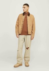 Nuori mies, jolla on päällään vaaleanruskea takki ruskean vetoketjupuseron päällä, beige revenneet farkut ja vaaleanruskeat kengät, seisoo yksinkertaisen vaalean taustan edessä.