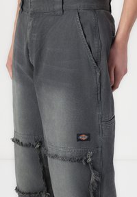 Gray denim pants with a distressed finish, featuring frayed edges, side pockets, and a small branded patch on the left leg.