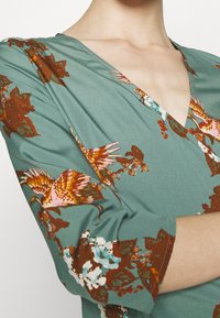 V-kauluspuku, jossa on lyhyet hihat, tehty teal-värisestä kankaasta, jossa on monivärinen kukka- ja lintukuosi. Ominaisuuksiin kuuluu virtaava muotoilu ja pehmeä rakenne.