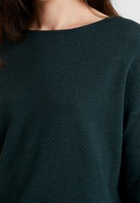 Detailní záběr osoby, která nosí tmavě zelený texturovaný pletený svetr s kulatým výstřihem a dlouhými rukávy.