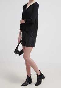 Zwarte blouse met lange mouwen en V-hals, gecombineerd met zwarte leren shorts en enkelboots met studs. Minimalistisch ontwerp.