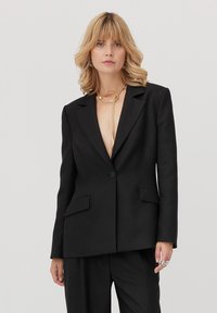 Czarna, dopasowana marynarka z klapą z wcięciem, dwoma przednimi kieszeniami, zamykana na jeden guzik, o luźnym kroju. Materiał wydaje się gładki i strukturalny.