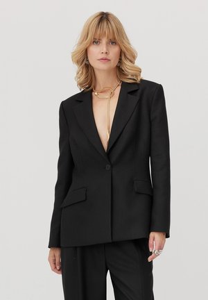 Czarna, dopasowana marynarka z klapą z wcięciem, dwoma przednimi kieszeniami, zamykana na jeden guzik, o luźnym kroju. Materiał wydaje się gładki i strukturalny.