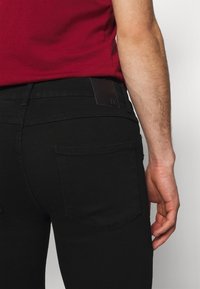 Jeans de mezclilla negra con un corte slim, que cuentan con un parche de cuero en la cintura y dos bolsillos traseros con detalles de costura sutiles.