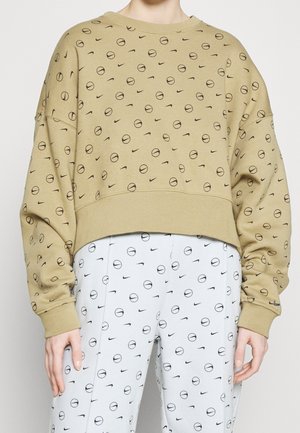 Person iført en beige sweatshirt og hvide bukser, begge med gentagne sorte abstrakte cirkulære og swoosh mønstre.