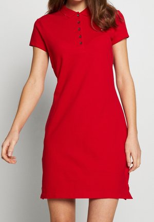 Mujer con un vestido polo rojo de manga corta y cuello con botones, de pie frente a un fondo liso de color gris claro.