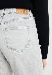 Jeans de denim cinza claro com um patch de couro da marca VMD, apresentando um estilo clássico de cinco bolsos e uma textura ligeiramente desbotada.