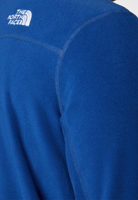 Blå fleecejacka med synliga sömmar och vit "THE NORTH FACE"-logotyp broderad på den övre ryggen nära axeln.