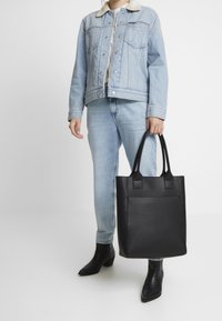 Mustat nahkainen olkalaukku, jossa on teksturoitu pinta ja kaksi yläkahvaa, jota kantaa henkilö vaaleansinisessä farkkutakissa ja farkuissa, mustissa korkokengissä.