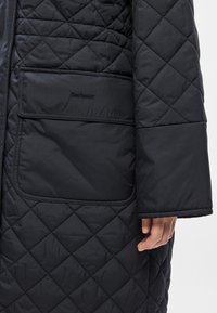 Μαύρο παλτό καπιτονέ με μοτίβο διαμαντιού, με τετράγωνη τσέπη με ανάγλυφο λογότυπο και φαρδιά, ίσια μανίκια. Λεία υφασμάτινη υφή.