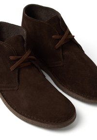 Botas de tobillo de ante marrón con punta redondeada, dos ojales y cordones marrones. El material texturizado contrasta con los bordes lisos y las costuras.