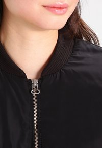 Närbild av en persons nedre ansikte och nacke som bär en svart jacka med en stor silverdragkedja och ribbad krage.