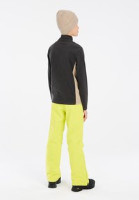 Schwarzer und brauner Fleece-Pullover, gelbe wasserdichte Hose und eine beigefarbene Strickmütze. Sichtbare Nähte und eine lässige Passform, geeignet für die Outdoornutzung.