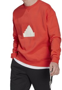 Person klädd i en röd sweatshirt med en vit geometrisk design på bröstet och svarta byxor med vita ränder på sidan.