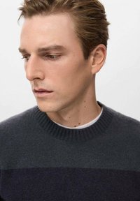 Giovane uomo con capelli castano chiaro che guarda in basso e di lato, indossa un maglione grigio scuro sopra una camicia bianca, sfondo neutro.
