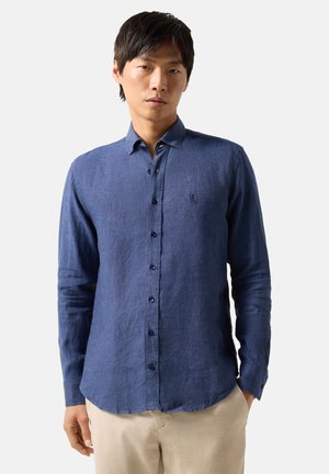 Männliches Model trägt ein langärmeliges blaues Hemd mit Knöpfen und beigefarbene Hose, steht vor einem einfarbigen hellen Hintergrund.