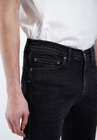 Los jeans de mezclilla negros presentan un ajuste slim, diseño de cinco bolsillos y herrajes plateados. La tela tiene un ligero brillo y costuras visibles.