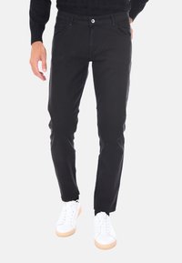 Uomo che indossa pantaloni neri attillati e sneakers bianche, con una mano rilassata al lato e l'altra in tasca.