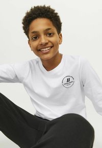 Vit långärmad T-shirt med en cirkulär svart logotyp med text på bröstet, ihop med svarta byxor, på en ljus bakgrund.