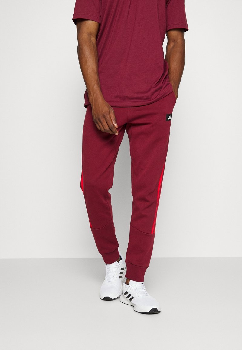 Homme portant un pantalon de jogging bordeaux avec des rayures rouges sur les côtés et des baskets blanches avec des rayures noires, se tenant devant un fond clair.
