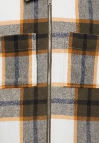 Plaid flannel jacket in white, grey, and brown with orange accents, featuring a zipper front and two chest pockets. Soft texture.