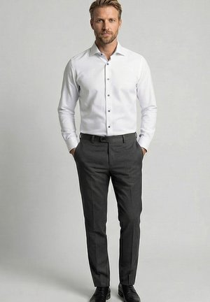 Man draagt een wit overhemd, donkere grijze pantalon, zwarte riem en zwarte nette schoenen, staat met handen in de zakken tegen een effen achtergrond.