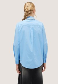 Cămașă button-up de culoare albastră deschisă, cu mâneci lungi, guler, detaliu cu pliu pe spate și manșete butonate. Țesătură de bumbac neted.