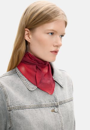 Młoda kobieta o prostych blond włosach nosi przezroczysty czerwony szalik na szyi i jasnoszarą kurtkę dżinsową, patrząc w prawo na białym tle.