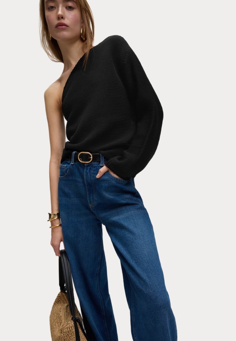 Un pull noir en maille, dégageant les épaules, associé à un jean bleu taille haute. Les accessoires comprennent une ceinture dorée, des bracelets et un sac en paille.