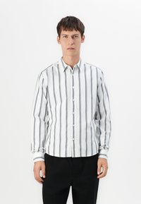 Joven vestido con una camisa blanca de manga larga y botones, con rayas verticales negras y pantalones negros, de pie frente a un fondo blanco.