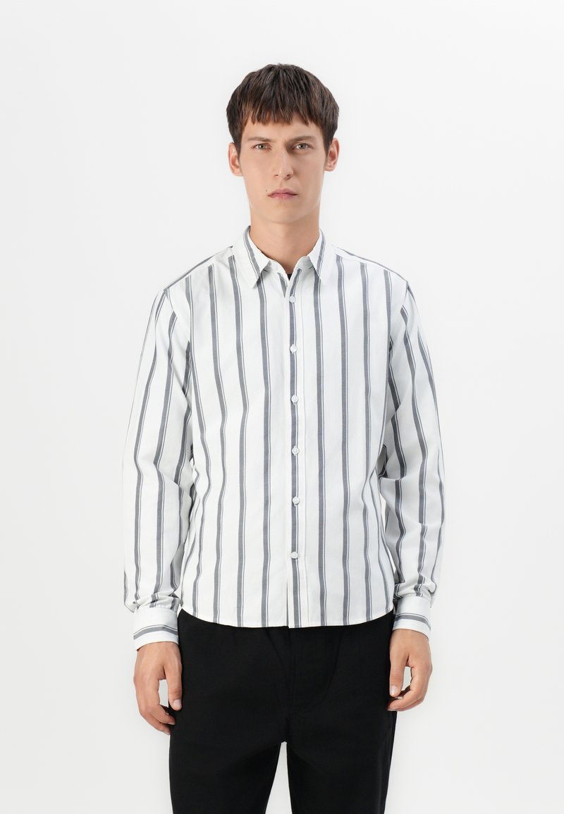Joven vestido con una camisa blanca de manga larga y botones, con rayas verticales negras y pantalones negros, de pie frente a un fondo blanco.