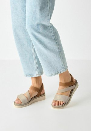 Infradito beige e marroni con suola imbottita, caratterizzati da più cinturini incrociati e una palette di colori neutrali. Indossati con jeans azzurri chiari.