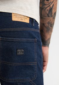 Uomo che indossa jeans di denim blu scuro con etichette del marchio visibili sulla tasca posteriore e in vita, che mostra un avambraccio tatuato.