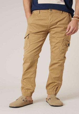 Homme portant un pantalon cargo beige avec des poignets élastiques et des chaussures beige à enfiler, debout les mains dans les poches devant un fond neutre.