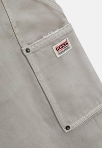 Tessuto grigio con una finitura testurizzata presenta una tasca a pattina e un'etichetta con dettagli rossi e neri, fissata da accessori neri.